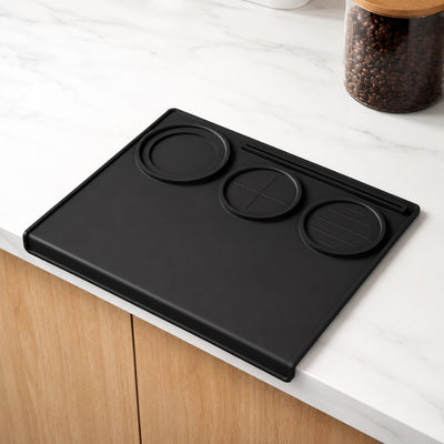 Black silicone espresso tamping mat on white kitchen counter beside coffee bean jar