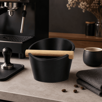 Matte charcoal black oval coffee knock box sitting next to a home espresso machine and coffee grinder.