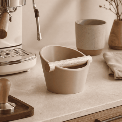 Desert Tan compact coffee knock box on a kitchen countertop alongside a ceramic mug and tamper.