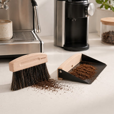 Coffee cleaning brush and scoop on a kitchen counter with coffee grounds, next to a coffee machine.