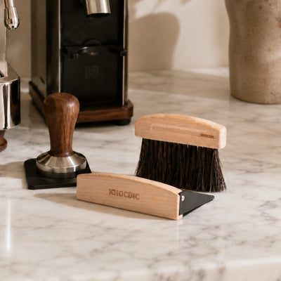 Wooden coffee brush and tampers on a marble surface with a coffee machine in the background.