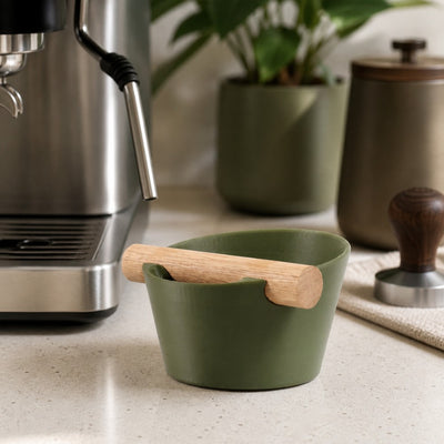 Hand tapping a portafilter against a matte green Nordic espresso knock box on a kitchen countertop.