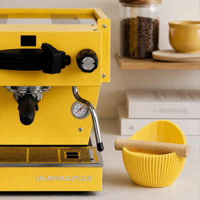 A matte yellow Nova coffee waste bin sitting beside a vibrant yellow home espresso machine and grinder setup.