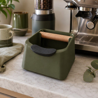 Green knock box with a wooden bar on a kitchen counter with coffee-making equipment in the background.