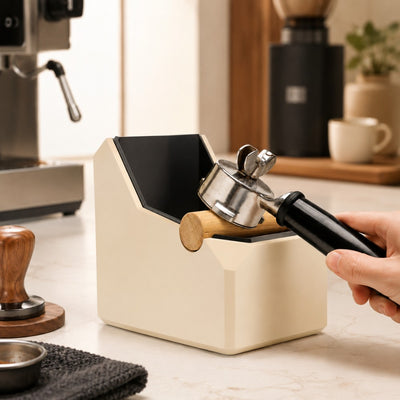 Hand knocking out an espresso puck using a portafilter over a tan rectangular coffee waste bin.