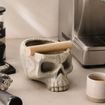 A metallic gold skull espresso knock box styled on a kitchen counter next to premium coffee beans.