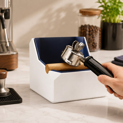 Rapping a portafilter to discharge a coffee puck into a large white dual-tone square knock box on a kitchen counter.