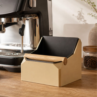 Latte Brown and black coffee knock box next to an espresso machine on a wooden surface.