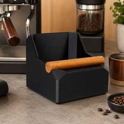 Black coffee waste bin with wooden handle on a kitchen counter with coffee beans and a coffee machine in the background.
