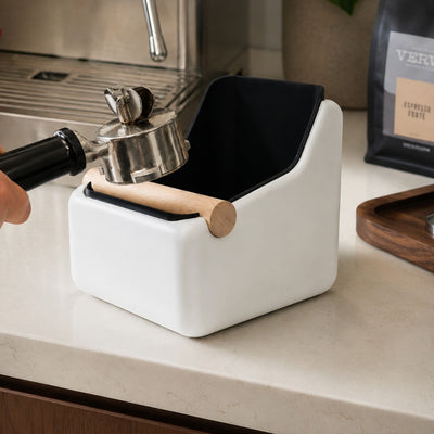 Emptying a spent coffee puck from an espresso portafilter into a large sculpted white and black square knock box.